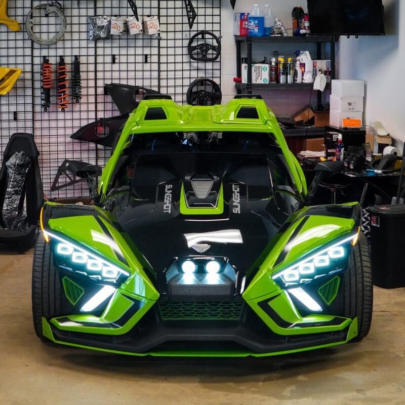A vibrant green and black Slingshot sports car with futuristic headlights in a workshop.