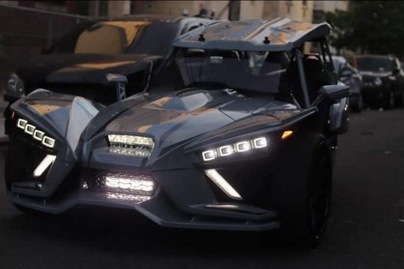 A sleek black three-wheeled vehicle with modern LED headlights on a street.