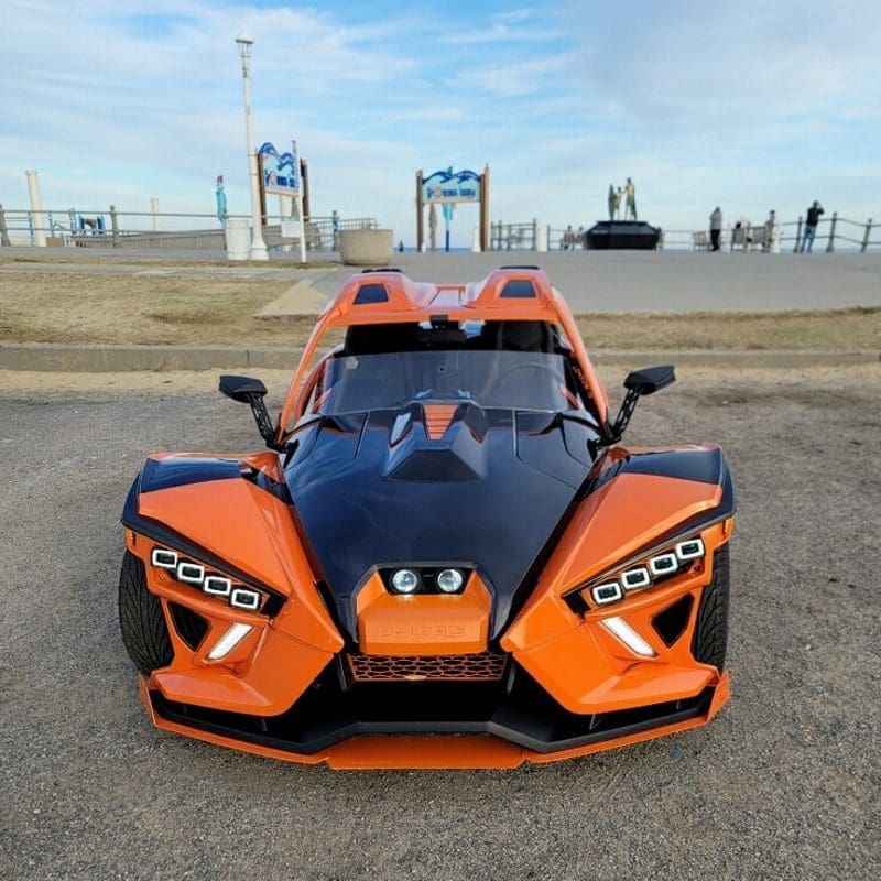Sleek orange and black Polaris three-wheeled vehicle parked on gravel.