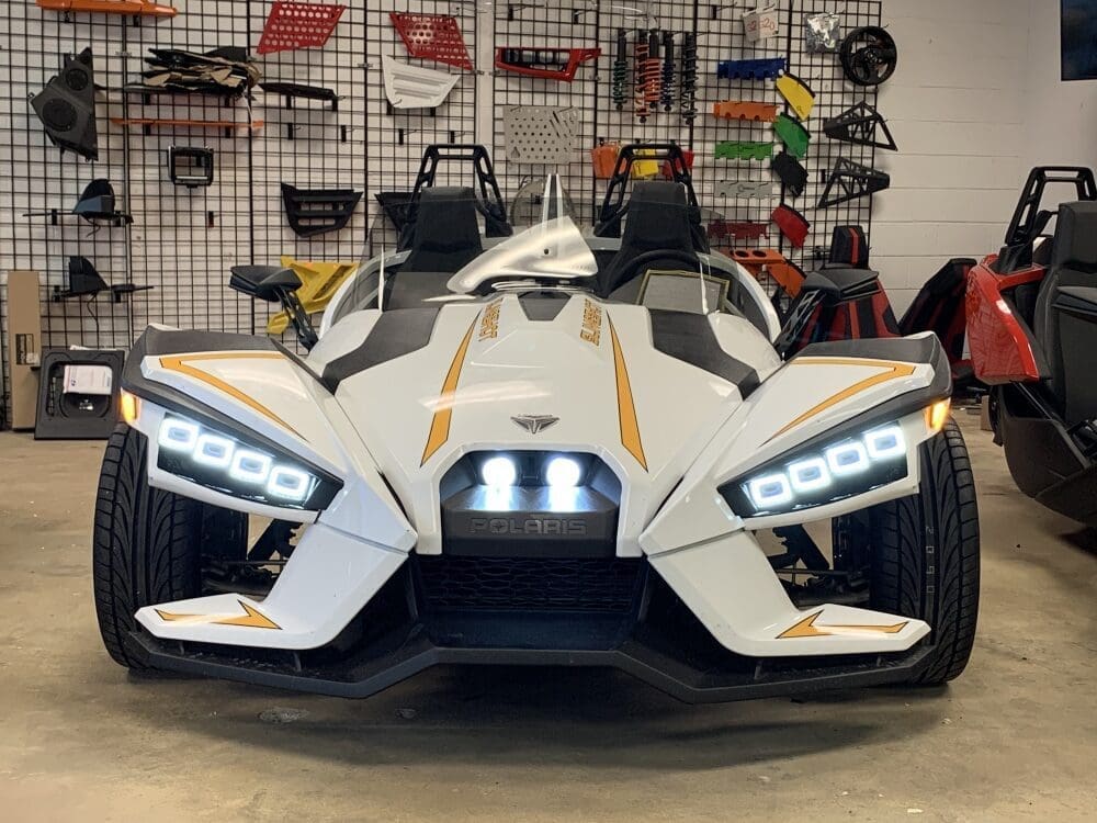 Front view of a sleek white Polaris three-wheeled vehicle with illuminated headlights.