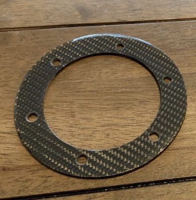 IMG_6980 Carbon fiber circular gasket with six holes on a wooden surface.