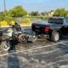 Pickup truck towing an ATV on a trailer in a parking lot.