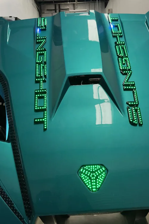 2594fc_3288ed35fc984f1e9022a19a985d92d6f002 Close-up of a teal sports car hood with green LED lights and a glowing emblem.